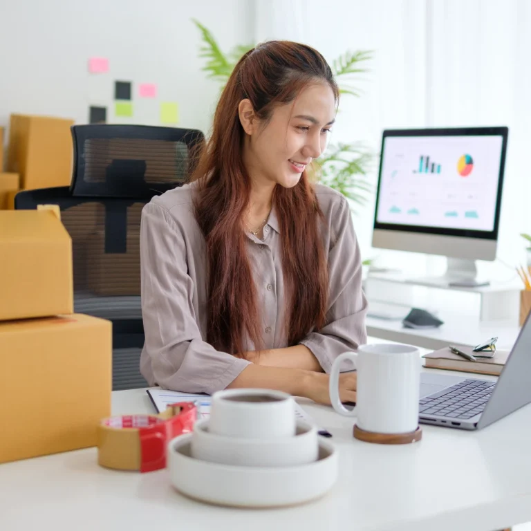 A cheerful young Asian woman with long brown hair, wearing a mauve shirt, smiles while working at a modern, bright desk.