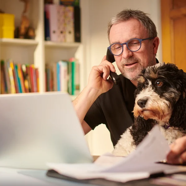 Man working on a laptop at home while talking on the phone, with a dog sitting beside him, representing a small or growing home-based business owner.