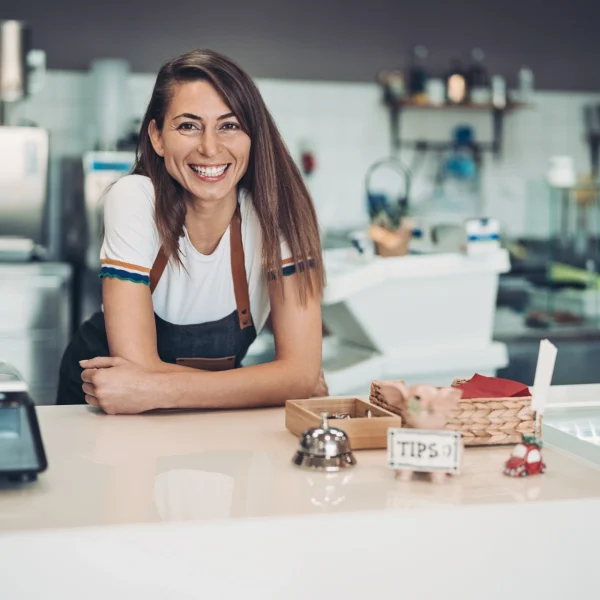 Smiling woman standing behind the counter of a small café, representing a small and growing business owner welcoming customers.